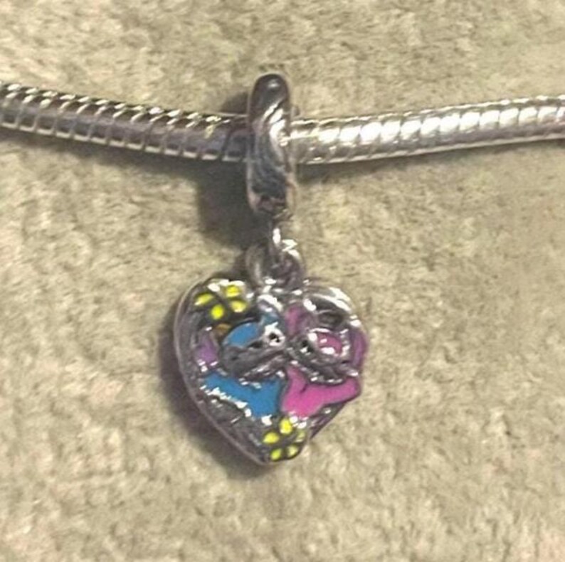 May include: A silver charm with a heart-shaped pendant featuring a blue and pink cartoon character design. The charm is attached to a silver chain. Yellow flowers are scattered around the characters. The charm is likely for a bracelet or necklace.