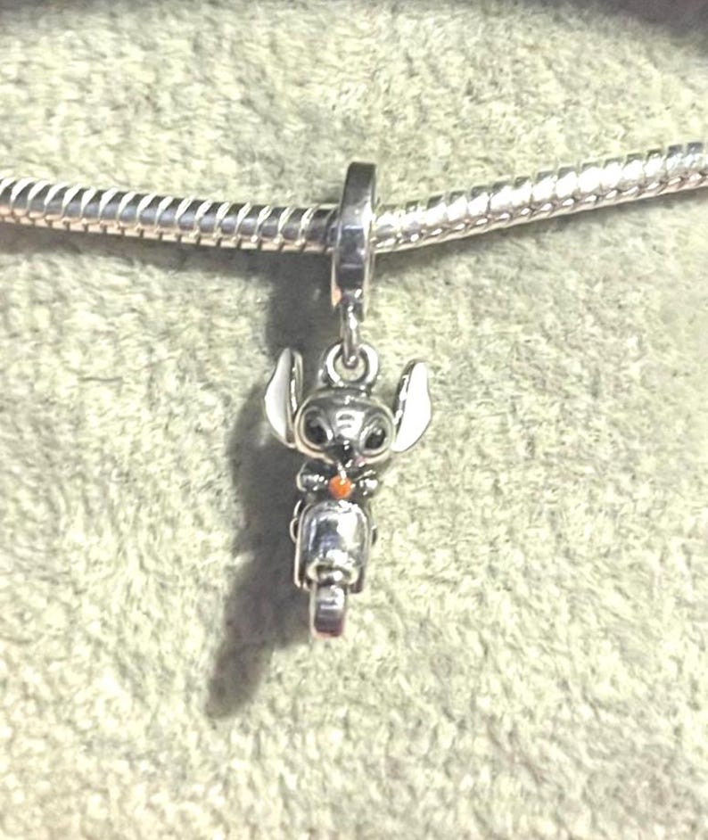 Puede incluir: Un charm de plata con el personaje Stitch de Lilo & Stitch en una scooter. El charm tiene detalles blancos y negros en las orejas y los ojos, y un acento naranja. Cuelga de una cadena de plata.
