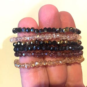 May include: A collection of beaded bracelets in various colors, including black, clear, pink, and gold. The bracelets are made of small, faceted beads, creating a sparkling effect. The bracelets are stacked together, showcasing the different colors and styles.