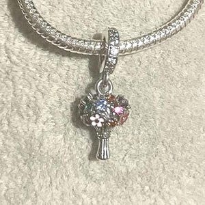 May include: A silver charm with a bouquet of colorful gemstones. The charm features a detailed flower arrangement with blue, pink, and white accents. The charm is attached to a silver bracelet.