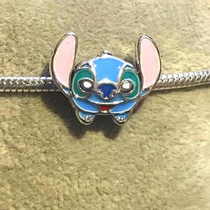 Może przedstawiać: Niebieski, zielony, r&oacute;żowy i srebrny charms z postacią Stitch z Lilo & Stitch. Charms ma szczeg&oacute;łowy design z uśmiechniętą twarzą, dużymi uszami i jest przymocowany do srebrnej bransoletki. Charms ma około 2,5 cm.