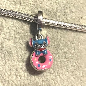 May include: A charm featuring a blue cartoon character with pink ears and a red collar, sitting atop a pink donut with blue sprinkles. The charm is attached to a silver bracelet. The charm is likely made of metal and enamel.