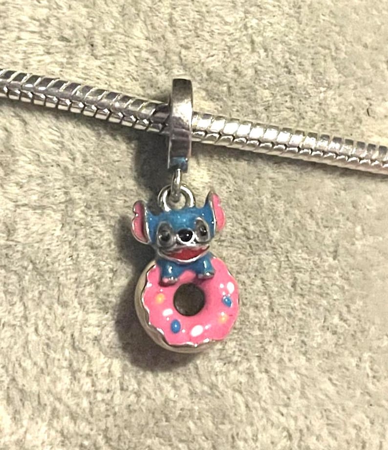 May include: A charm featuring a blue cartoon character with pink ears and a red collar, sitting atop a pink donut with blue sprinkles. The charm is attached to a silver bracelet. The charm is likely made of metal and enamel.