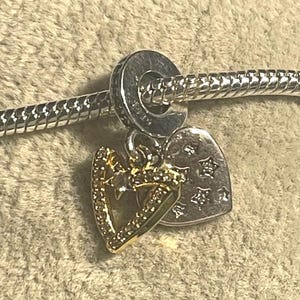 May include: A silver charm with a gold-colored heart-shaped pendant, encrusted with small clear stones, and a silver heart-shaped pendant with star cutouts. The charm is attached to a silver bracelet.