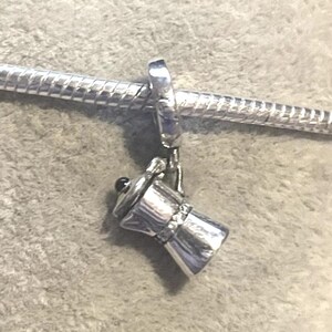 May include: A silver charm featuring a miniature coffee maker, suspended from a silver chain. The charm has a detailed design, with a small handle and a black knob on top. The charm is designed to be a pendant or a bracelet charm.