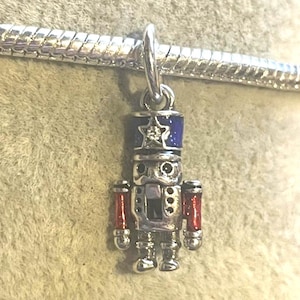 May include: A silver charm in the shape of a nutcracker soldier. The charm has a blue hat with a star, red arms, and detailed silver features. It is attached to a silver bracelet.