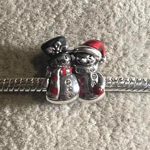 May include: A silver charm featuring two snowmen. One snowman wears a black top hat and red scarf, the other a red Santa hat and scarf. Both have silver bodies and are attached to a silver bracelet.