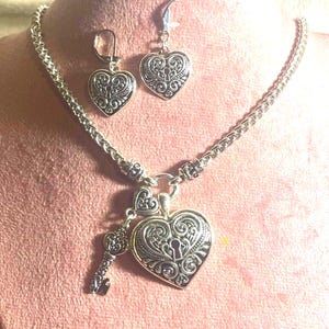 May include: A silver-toned jewelry set featuring a necklace with a heart-shaped pendant and key charm, plus matching heart earrings. The heart pendants and earrings have an ornate, filigree design. The jewelry is displayed on a pink surface.