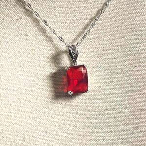 May include: A silver-toned necklace featuring a rectangular, faceted red gemstone pendant. The pendant is held by a silver-toned metal bail attached to a delicate, twisted silver-toned chain. The design is simple, focusing on the vibrant red stone.