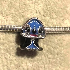 May include: A blue and black charm featuring a cartoon character with large black eyes and a small red mouth. The charm is on a silver bracelet. The charm has a blue head and black body.