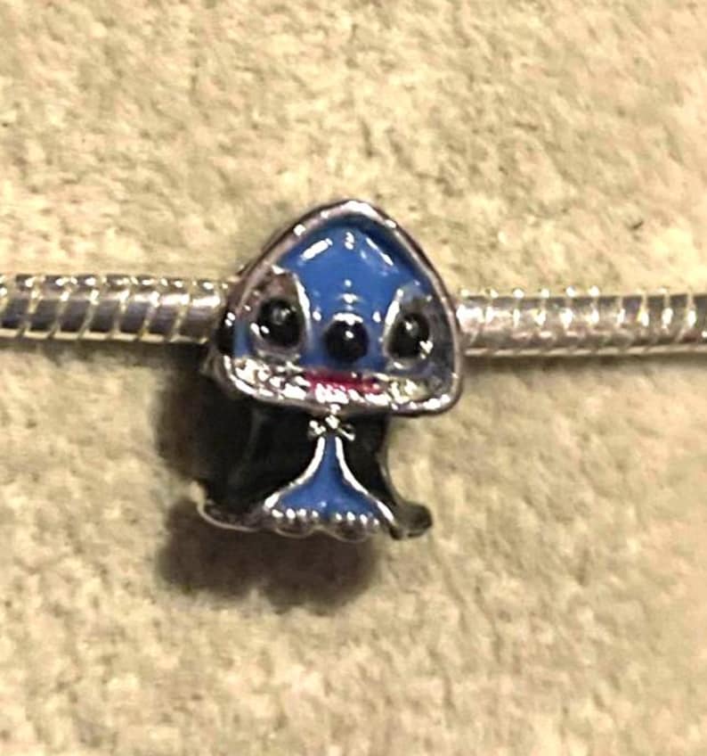 May include: A blue and black charm featuring a cartoon character with large black eyes and a small red mouth. The charm is on a silver bracelet. The charm has a blue head and black body.