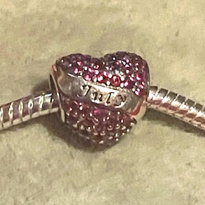 May include: A heart-shaped charm with a silver band and a surface covered in small, sparkling pink stones. The word "Juicy" is engraved on the silver band. The charm is attached to a silver bracelet.