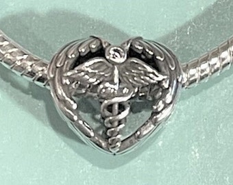 May include: A silver charm with a heart-shaped frame. Inside the heart is a medical symbol with wings and a serpent-entwined staff. The charm is attached to a silver bracelet. The background is a light teal color.