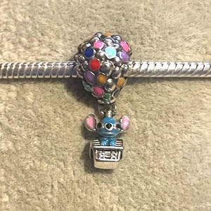 Może przedstawiać: Urok z postacią Stitcha z Lilo & Stitch w balonie na ogrzane powietrze. Balon wykonany jest z kolorowych koralik&oacute;w, a Stitch znajduje się w srebrnym koszu. Urok jest przymocowany do srebrnej bransoletki.