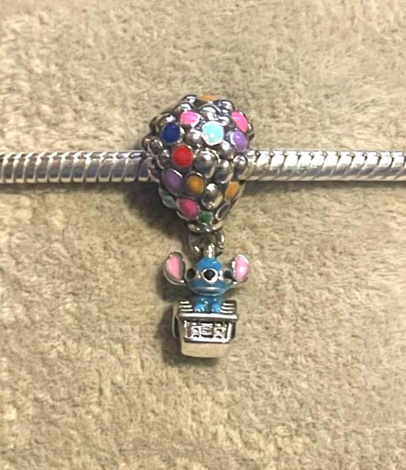 Może przedstawiać: Urok z postacią Stitcha z Lilo & Stitch w balonie na ogrzane powietrze. Balon wykonany jest z kolorowych koralik&oacute;w, a Stitch znajduje się w srebrnym koszu. Urok jest przymocowany do srebrnej bransoletki.