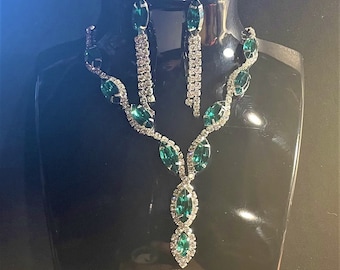 May include: A dazzling emerald green and silver necklace and earring set. The necklace features marquise-cut green stones and sparkling clear accents. The matching earrings have a dangling design. The jewelry is displayed on a black mannequin.