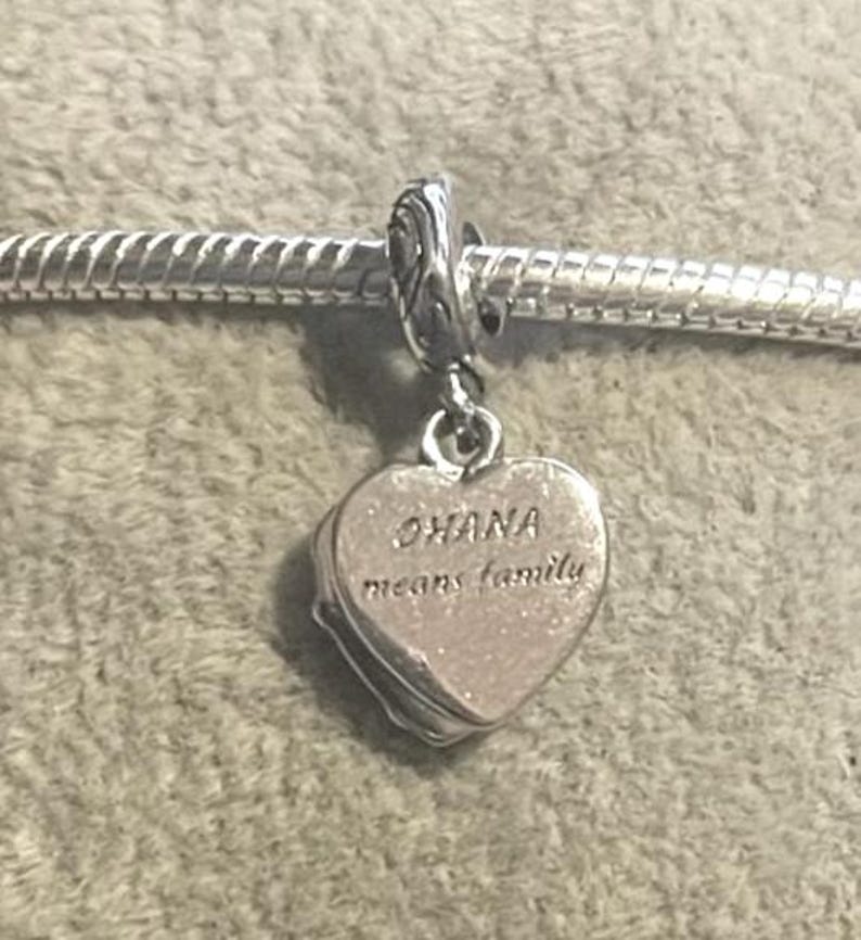May include: A silver charm with a heart-shaped pendant. The pendant has the words "OHANA means family" engraved on it. The charm is attached to a silver bracelet.
