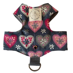 May include: A cat harness with a heart pattern in pink, gray, and white on a dark gray background. The harness has a D-ring for leash attachment and a label that reads "Rose & Bramble Cat Harness".