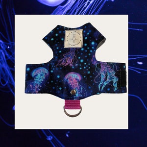 Peut inclure: Harnais pour chat avec un motif de méduses bleues, violettes et roses sur fond bleu foncé. Il est équipé d'une sangle rose et d'un anneau métallique. Le texte "ROSE & BRAMBLE CAT HARNESS" est imprimé. Conçu pour les chats.