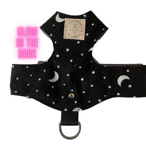 May include: Black cat harness with a white star and crescent moon pattern. The harness has a metal D-ring and a small label with a cat silhouette. The text "Glow in the Dark" is visible in the image.