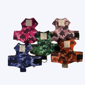 May include: Five dog harnesses in camouflage patterns: pink, green, brown, blue, and orange. Each harness features a rectangular label with a logo and adjustable straps with Velcro fastenings. A metal D-ring is included for leash attachment.