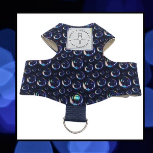 May include: A cat harness featuring a navy blue pattern of iridescent bubbles. The harness includes a white label with the text "ROSE & BRAMBLE CAT HARNESS" and a cat silhouette. A navy strap with a silver D-ring is attached.