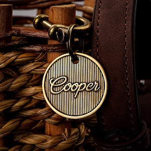 May include: A round, gold-colored pet tag with the name "Cooper" in elegant script. The tag has vertical line detailing and hangs from a bronze-colored clasp. A brown leather collar is visible in the background, along with woven wood.