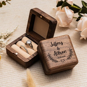 May include: A wooden ring box with a lid that opens to reveal a velvet interior with two ring slots. The box is engraved with the names "Jeffrey & Allison" and the date "07.09.2024".