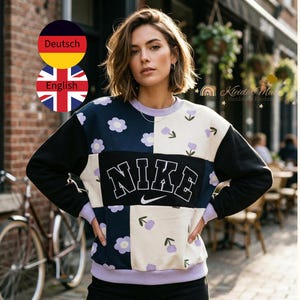 Könnte beinhalten: Ein Sweatshirt von Nike im Patchwork-Stil mit dem Markenlogo in Weiß. Das Design umfasst schwarze, marinefarbene und cremefarbene Paneele, die mit lila Blumenmustern verziert sind. Es hat einen lila Kragen und Manschetten.