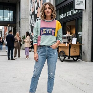 May include: A woman wearing a color-block sweatshirt with the Levi's logo and light blue jeans. The sweatshirt features pastel green, pink, and yellow panels. She is wearing white sneakers. The background includes a building with a mural and a sign that says "Urban Cultivate."