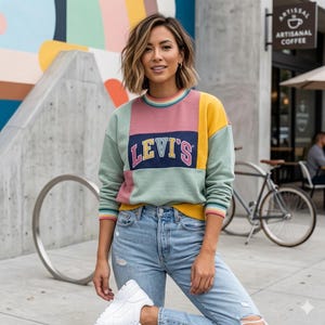 May include: A woman wearing a color-block Levi's sweatshirt with a navy blue rectangle featuring the brand's logo. The sweatshirt has panels of pink, yellow, and mint green. She is wearing light blue ripped jeans and white sneakers.