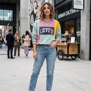 Pode incluir: Uma mulher usando um moletom Levi's com blocos de cores e jeans azul claro. O moletom apresenta pain&eacute;is em verde pastel, rosa e amarelo. Ela est&aacute; usando t&ecirc;nis brancos. O fundo inclui um pr&eacute;dio com um mural e uma cafeteria.