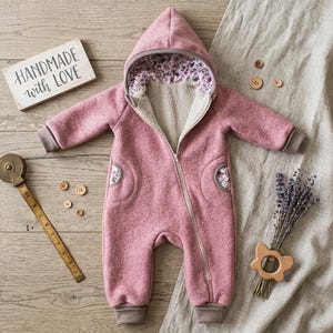 May include: A dusty pink hooded jumpsuit featuring a diagonal front zipper, beige fleece lining, and floral patterned accents inside the hood and pockets. The garment is displayed alongside a wooden sign reading HANDMADE with LOVE, buttons, and a measuring tape.