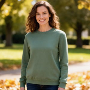 Pode incluir: Uma mulher vestindo um moletom verde s&aacute;lvia com gola redonda. O moletom tem mangas compridas e punhos e bainha canelados. A mulher est&aacute; sorrindo e em p&eacute; ao ar livre em um parque com &aacute;rvores e folhas ca&iacute;das.
