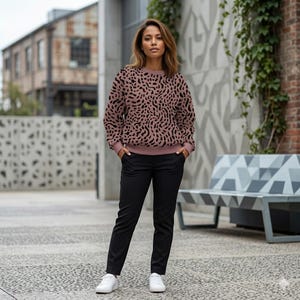 May include: A person wearing a mauve-colored sweatshirt with a black, abstract pattern, paired with black pants and white sneakers. The sweatshirt has a crew neck and long sleeves. The outfit is suitable for casual wear.