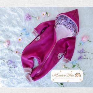Può includere: Una tutina con cappuccio color magenta con chiusura a zip frontale, maniche lunghe e polsini a coste rosa chiaro. L'interno del cappuccio presenta un motivo floreale. Un'etichetta bianca riporta il testo KleiderMai Handmade Kleidung.