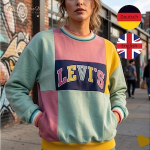 Könnte beinhalten: Ein Sweatshirt mit Farbblock-Design und dem Levi's-Logo. Das Sweatshirt hat ein Patchwork-Design mit rosa, gelben und mintgrünen Panels. Kragen und Manschetten haben Regenbogenstreifen. Das Model steht vor einer Backsteinmauer.