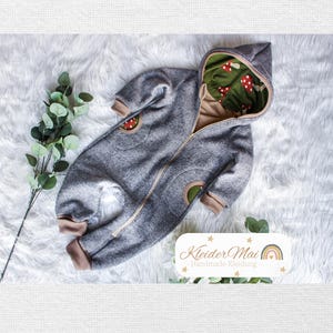 Op de afbeelding: Een grijze jumpsuit met capuchon, ritssluiting aan de voorzijde, beige boorden en twee zakken. De binnenkant van de capuchon is gevoerd met groene stof met een patroon van rode en witte paddenstoelen. Een label toont Kleider Mai Handmade Kleidung.