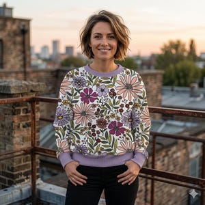 May include: A floral print sweatshirt featuring a white background with large pink, purple, and yellow flowers. The sweatshirt has a lilac collar and cuffs. The person is wearing black pants.