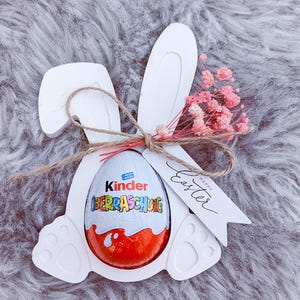 Könnte beinhalten: Weißer Holzschnitt eines Hasen mit einem Kinder Überraschungsei darin. Der Hase hat ein Schild mit der Aufschrift "Frohe Ostern".