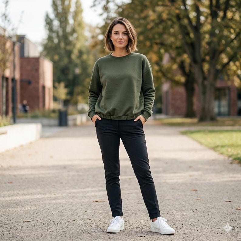 Pode incluir: Uma mulher vestindo um moletom verde-oliva com cintura ajustada, cal&ccedil;as pretas e t&ecirc;nis brancos. A roupa &eacute; casual e adequada para atividades ao ar livre. O fundo apresenta uma passarela pavimentada e &aacute;rvores.