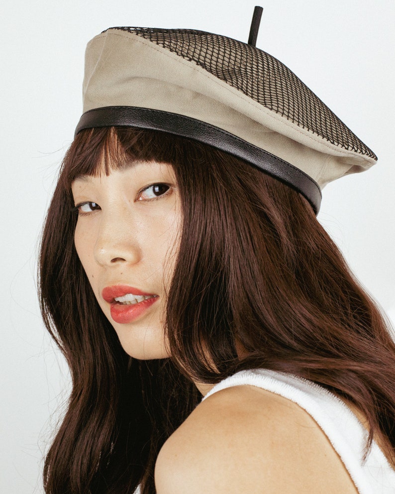 May include: A woman wearing a beige beret with a black mesh panel and a black leather band. The beret has a small black antenna on top.