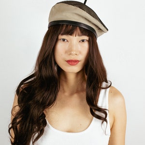 May include: A woman with long brown hair is wearing a beige beret with a black mesh panel and a black leather band. The beret has a small black straw sticking out of the top.