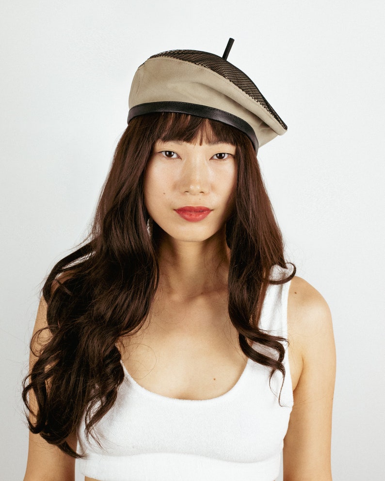 May include: A woman with long brown hair is wearing a beige beret with a black mesh panel and a black leather band. The beret has a small black straw sticking out of the top.
