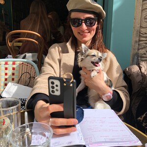 May include: A woman wearing a beige beret and sunglasses holds a small white and brown chihuahua dog. She is sitting at a table with a black phone case with a ring attached, a glass of water, and a notebook with handwritten notes.