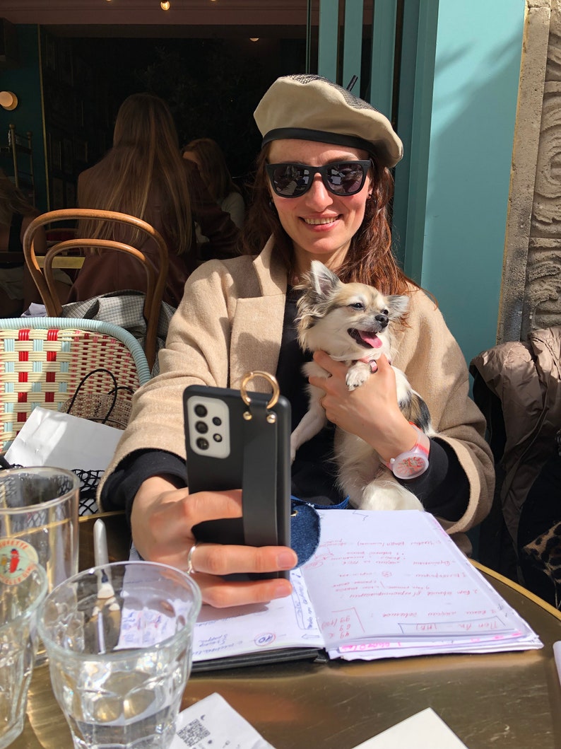 May include: A woman wearing a beige beret and sunglasses holds a small white and brown chihuahua dog. She is sitting at a table with a black phone case with a ring attached, a glass of water, and a notebook with handwritten notes.