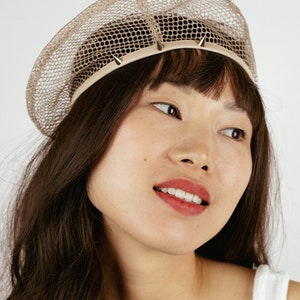 May include: A woman with long brown hair is wearing a beige mesh beret with silver studs around the brim. The beret is a unique and stylish accessory.