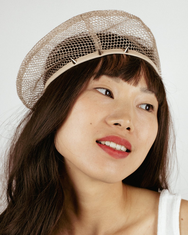 May include: A woman with long brown hair is wearing a beige mesh beret with silver studs around the brim. The beret is a unique and stylish accessory.