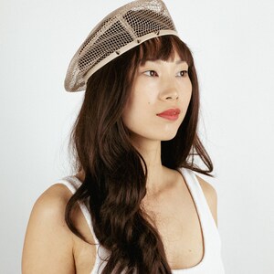 May include: A beige mesh beret with a decorative trim. The beret is worn by a person with long brown hair.