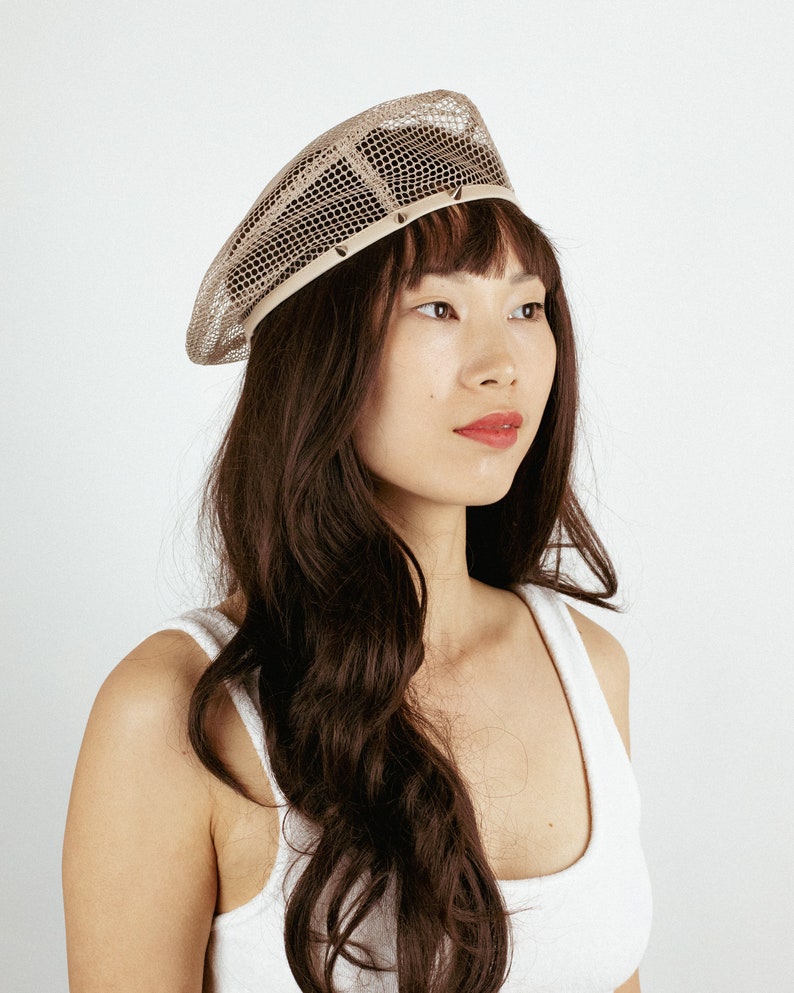 May include: A beige mesh beret with a decorative trim. The beret is worn by a person with long brown hair.